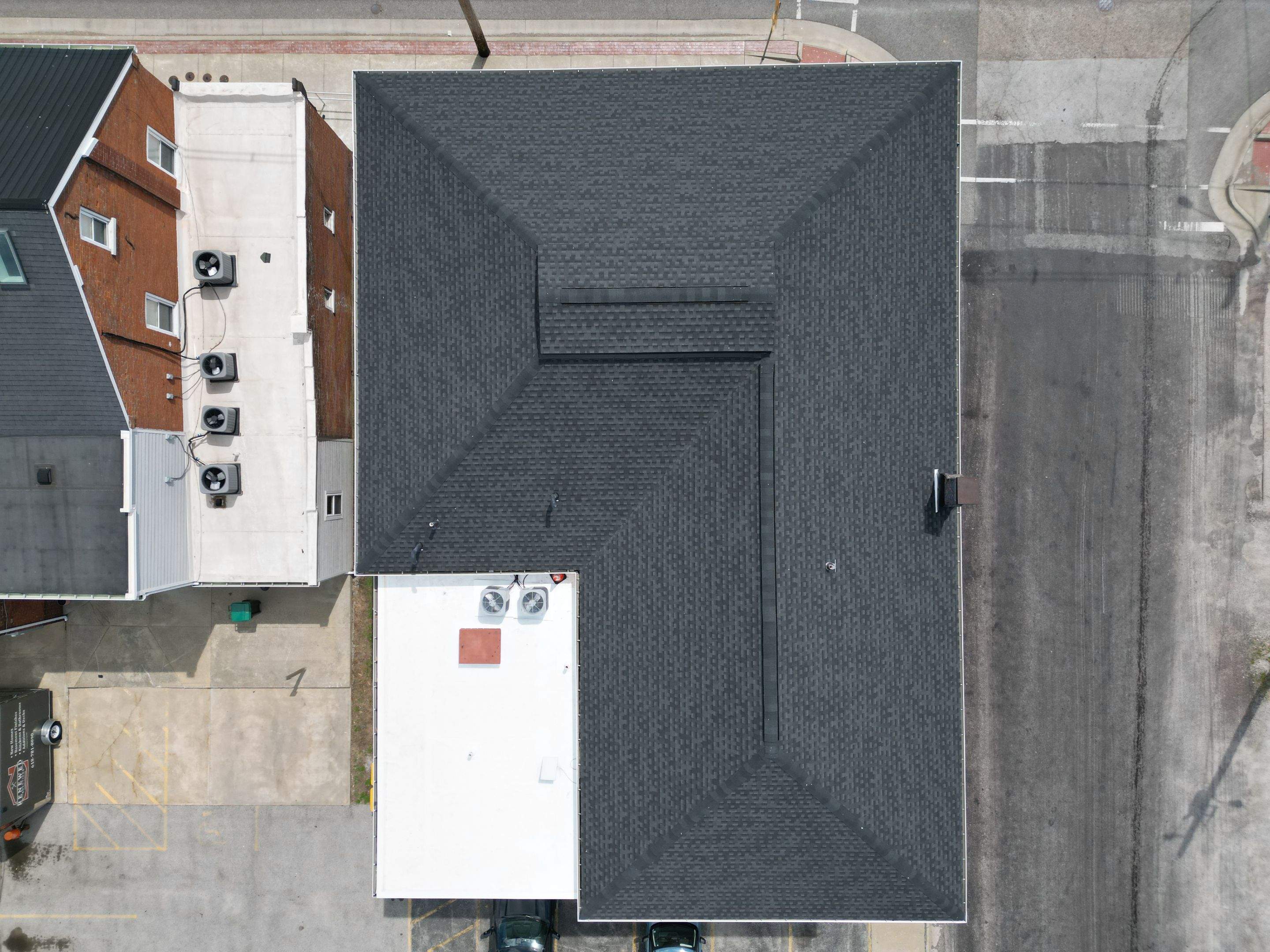 Aerial view of a completed dark architectural shingle hip roof on a commercial building in Millstadt, IL, alongside the adjoining flat membrane roof section with rooftop units