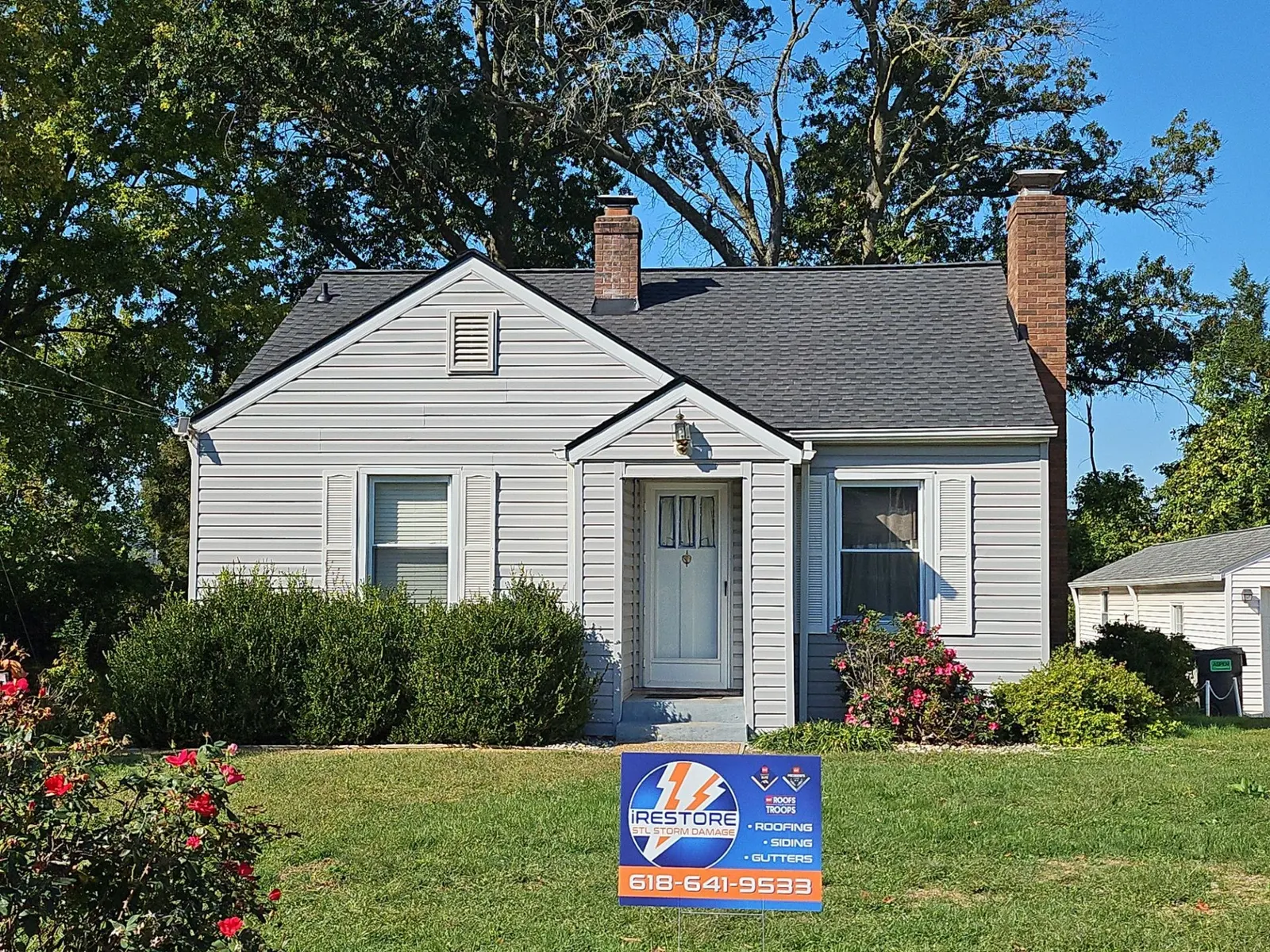 Newly finished charcoal architectural shingle roof on a white-sided Cape Cod home with two brick chimneys in Belleville, IL, with an iRestore STL yard sign out front
