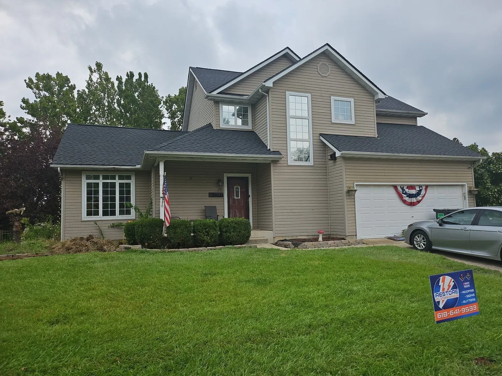 New charcoal architectural shingle roof on a tan-sided two-story home in Fairview Heights, IL, with an iRestore STL yard sign in the front lawn