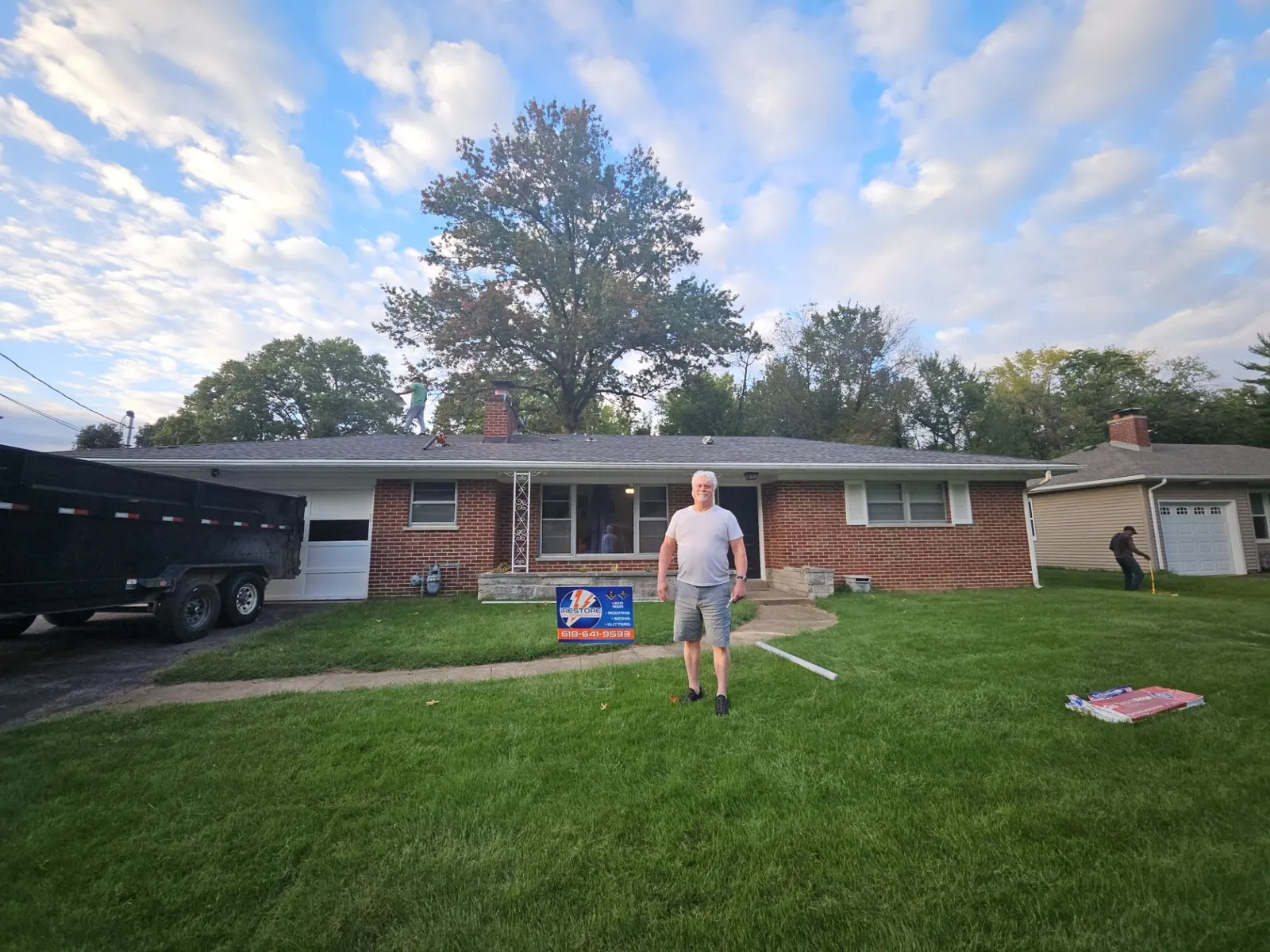 Smiling homeowner standing in his front yard in Swansea, IL, in front of his brick ranch home and its newly finished roof, with an iRestore STL yard sign
